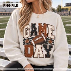 Op de afbeelding: Een crèmekleurige sweater met ronde hals en de tekst GAME DAY in een verweerd bloklettertype. Het ontwerp bevat een bruine football-afbeelding in het midden tussen de woorden, geflankeerd door twee kleine sterren en horizontale strepen.