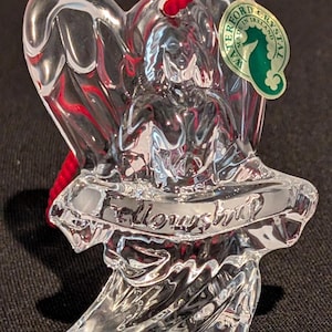 Op de afbeelding: Transparante kristallen engelornament met een rood koord om op te hangen. Het ornament heeft het woord "Fellowship" erin gegraveerd. Een groen en wit label met een zeepaardlogo leest "Waterford Crystal Made in Ireland."
