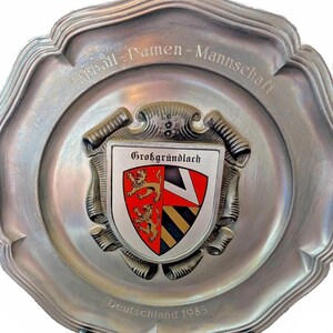 Könnte beinhalten: Eine silberfarbene Zierplatte mit gewelltem Rand. Die Platte trägt die Aufschrift "Fußball - Damen - Mannschaft" und "Deutschland 1985". Ein zentrales Emblem zeigt ein Schild mit einem roten und goldenen Löwen, einem schwarz-weißen Design und dem Text "Großgründlach".