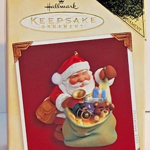 Könnte beinhalten: Hallmark Keepsake Weihnachtsschmuck in einer Box. Der Schmuck zeigt den Weihnachtsmann mit einem Sack voller Spielzeug. Auf der Box steht "Santa's Magic Sack" und "Clips to Light".