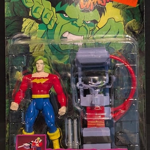 Könnte beinhalten: Doc Samson Actionfigur in einer Kunststoffverpackung. Die Figur hat grüne Haare, ein rotes Hemd, blaue Hosen und gelbe Stiefel. Die Verpackung enthält einen Omega Blaster mit "Missile-Firing"-Funktion und den Text "The Incredible Hulk".