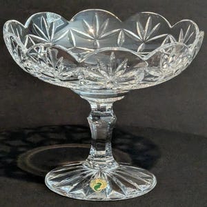 Puede incluir: Un cuenco de pedestal de cristal tallado transparente, con borde festoneado y patrones de estrella. El cuenco se asienta sobre un tallo facetado y una base circular. Una pegatina verde y dorada es visible en la base.