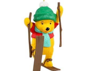May include: A festive Christmas ornament featuring Winnie the Pooh. He is depicted in a green hat with a white bobble, a red jacket, and a blue scarf, holding ski poles and wearing skis. The ornament is designed for hanging.