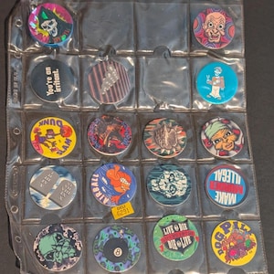 May include: A collection of vintage pogs in a clear plastic sheet. The pogs feature various colorful designs, including cartoon characters, text, and abstract patterns. Some pogs have text that reads "Dunk Slam", "Airball", and "Make Illegal".
