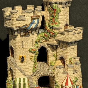 Può includere: Un castello in miniatura dettagliato con mattoni beige, con più torri, porte ad arco e una scala a chiocciola. Uno stendardo blu e bianco pende da una torre. Viti verdi con bacche rosse adornano le pareti e una tenda a strisce rosse e bianche è alla base.