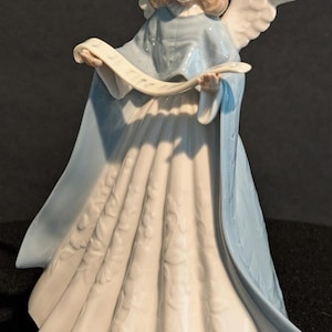 May include: A porcelain angel figurine with a serene expression, wearing a flowing blue and white gown. The angel holds a scroll and has detailed wings. The figurine is approximately 25 cm tall.