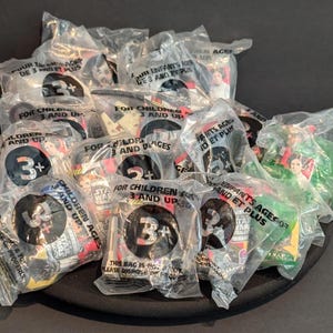 Pode incluir: Uma coleção de brinquedos Star Wars selados em sacos plásticos transparentes, cada um rotulado "Para crianças a partir de 3 anos". Os sacos apresentam imagens de personagens e o logotipo Star Wars.