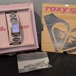May include: A silver-toned rectangular wristwatch with a light purple face, housed in a light pink box labeled Roxy Diamond Collection. The set includes a wooden lid with a large Roxy logo and a small plastic bag containing extra metal watch links.