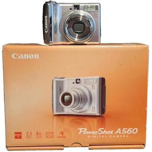 May include: A silver Canon PowerShot A560 digital camera is displayed on top of its original box. The box is orange with the Canon logo and a picture of the camera. The camera has a 4x zoom and 7.1 megapixels.