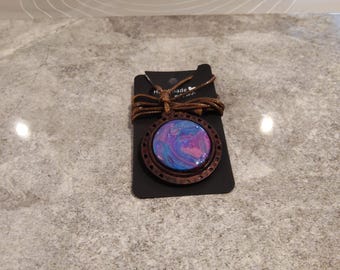 Puede incluir: Un collar con colgante hecho a mano con un diseño circular veteado en tonos azules, morados y rosas, enmarcado en un marco de madera marrón oscuro. El colgante cuelga de un cordón marrón, presentado en una tarjeta negra.