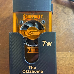 Puede incluir: Un dispositivo electrónico naranja y negro con el texto "ONEFINITY" y "J TECH Photonics". El dispositivo está etiquetado como "7W" y "7W+". La parte inferior del dispositivo tiene el texto "The Oklahoma Outdoorsman".