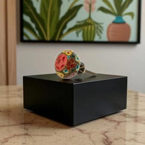 May include: A decorative cabinet knob with a floral design. The knob features a colorful pattern of red, yellow, and blue flowers on a clear, rounded surface. The knob is mounted on a black square base.