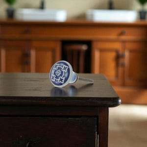May include: A blue and white ceramic cabinet knob with a floral design. The knob is round with a silver screw and is set against a dark wood surface. This decorative hardware is suitable for drawers and cabinets.