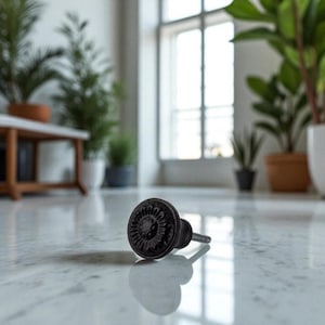 May include: A black, decorative cabinet knob with a floral design. The knob is made of metal and has a round shape. The knob is lying on a white marble surface, reflecting the light.
