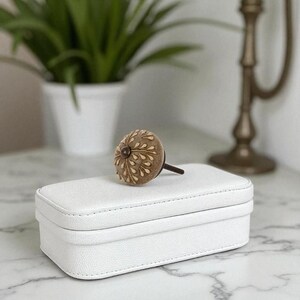 May include: A white rectangular jewelry box with a decorative brown knob. The knob has a floral design. The box is on a marble surface, with a plant and a decorative candle holder in the background.