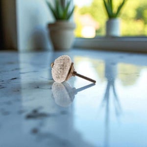 May include: A decorative cabinet knob with a textured, off-white, round head and a brass-colored base. The threaded screw extends from the back. The knob is resting on a reflective surface.