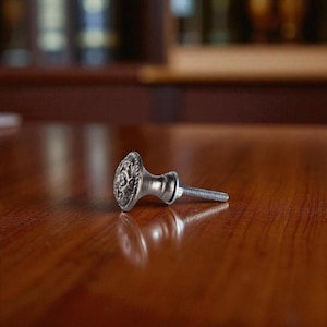 May include: A silver-toned cabinet knob with an ornate, floral design on the face. The knob is lying on a polished, dark wood surface. The screw is attached and extends to the right.