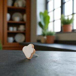 May include: A single, white, flower-shaped earring with a gold-colored edge and a silver post. The earring is set against a dark gray surface, with a blurred background featuring a wooden cabinet and a window with plants.