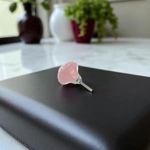 May include: A pink, decorative cabinet knob with a scalloped edge. The knob has a silver-colored stem and is set against a dark, textured surface. The background includes blurred greenery and vases.