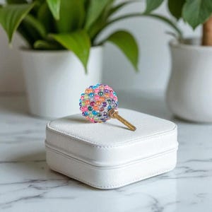 May include: A decorative bottle stopper with a colorful beaded sphere design. The sphere is composed of various colored beads and is attached to a gold-colored metal screw. The stopper rests on a white, square jewelry box.