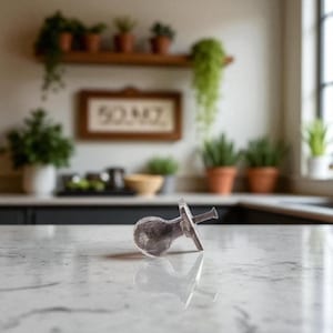 May include: A clear, glass cabinet knob with a silver-colored screw and a decorative, ridged edge. The knob is lying on a white countertop, reflecting its shape. The background includes a kitchen setting with plants and a framed sign.