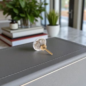 May include: A clear, decorative cabinet knob with a gold-colored screw and a semi-transparent, rounded head filled with small, light-colored stones. The knob is set against a gray surface, suggesting a piece of furniture or a drawer.