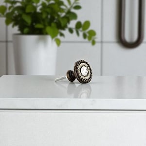 May include: A decorative cabinet knob with a bronze finish and a circular design. The knob features a central clear crystal surrounded by smaller crystals and a beaded outer edge. The hardware is designed for furniture and home decor.