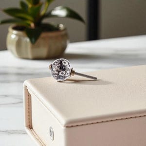 May include: A decorative cabinet knob with a white and black floral design. The knob is round and made of ceramic, with a metal screw. It rests on a light beige box. A small potted plant is in the background.