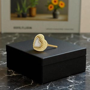 May include: A heart-shaped decorative object with a white heart-shaped center and a gold-colored border, resting on a black box. The object has a wooden stick attached to it. The background includes a blurred image of plants and flowers.