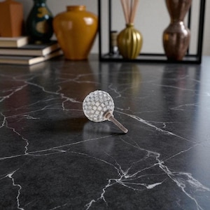 May include: A decorative cabinet knob with a mosaic-style, spherical design. The knob features a silver and white color scheme and a textured surface. The knob is set against a dark, marble-like surface, with other decorative items in the background.
