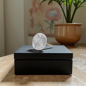 May include: A black box with a white and gray marble-patterned knob. The knob is oval-shaped and has a light-colored stem. The box is sitting on a wooden surface, with a painting and a potted plant in the background.