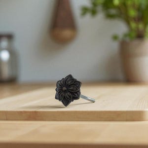 May include: A dark gray, flower-shaped cabinet knob. The knob has a detailed floral design and a silver-colored screw. The knob is on a light brown wooden surface.
