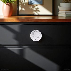 May include: A close-up of a black dresser drawer with a white, textured knob. The knob is round with a floral-like pattern. The drawer is part of a larger piece of furniture with a wooden top.