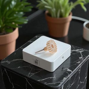 May include: A decorative cabinet knob with a clear, ridged, amber-colored design. The knob is mounted on a white square base. The base sits on a black and white marble-patterned surface. Small potted plants are in the background.