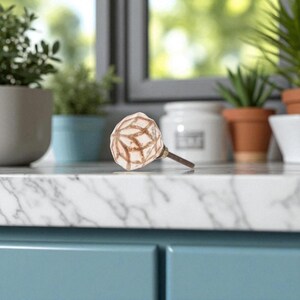 May include: A decorative cabinet knob with a cream-colored, geometric patterned ceramic top and a bronze-colored metal stem. The knob is set against a blurred background of plants and a window.
