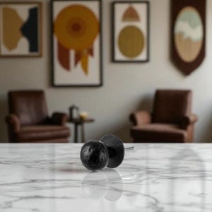 May include: A pair of black, textured cabinet knobs with a round, spherical design. The knobs are made of a dark material and are set against a white marble surface. The background features framed artwork and leather armchairs.