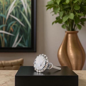 May include: A clear crystal cabinet knob with a silver-colored trim and a clear stem. The knob is round and sits on a black surface. The background includes a painting of green plants and a gold-colored vase.