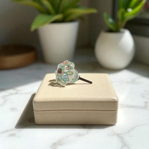 May include: A decorative cabinet knob with a floral design in shades of blue, green, yellow, and red. The knob is set on a beige square box. The knob has a dark brown stem and a round, light brown base.