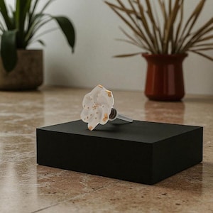 May include: A white and gold floral bottle stopper with a silver stem rests on a black rectangular block. The stopper has a flower design with gold accents. The background includes plants in pots.