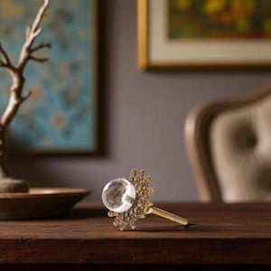 May include: A decorative cabinet knob with a clear, spherical glass center and an ornate, antique brass-colored base. The knob is set against a dark wooden surface, showcasing its intricate design and vintage aesthetic.