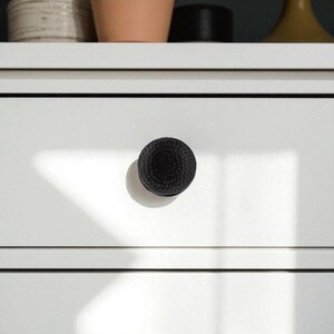 May include: Close-up of a white drawer with a black, textured knob. The knob has a woven rope design. The drawer is part of a white cabinet or dresser. The image is well-lit, highlighting the details of the knob and the drawer's surface.