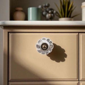 May include: A clear, flower-shaped cabinet knob with a bronze-colored center is mounted on a beige drawer. The knob's design includes a textured, frosted glass appearance. The drawer is part of a larger cabinet.
