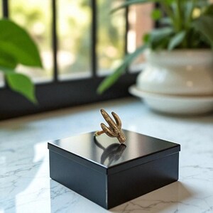 May include: A gold-colored cactus ring sits atop a black square box. The ring's design mimics the shape of a cactus. The box is on a white marble surface, with a blurred background of a window and plants.