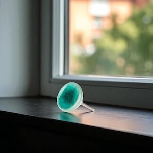 May include: A decorative glass cabinet knob with a clear, light green outer layer and a darker teal center. The knob has a small, clear stem and is set against a window background.