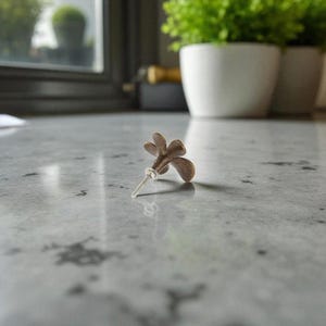 May include: A bronze-colored, flower-shaped cabinet knob with a clear stem. The knob is resting on a gray, speckled surface. The background includes a window and potted plants.