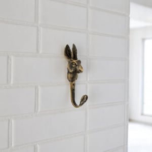May include: A bronze-colored wall hook shaped like a rabbit's head. The hook is mounted on a white tiled wall. The rabbit's head is detailed with textured fur and a downward-facing hook.