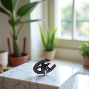 May include: A black, cast-iron cabinet pull in the shape of a horseshoe. The pull is mounted on a marble surface, with a screw visible. The background includes potted plants and a window.