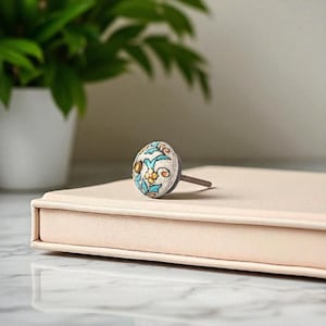 May include: A decorative ceramic cabinet knob with a floral design in teal, yellow, and brown on a cream background. The knob is mounted on a threaded metal screw. The knob is resting on a light pink surface.