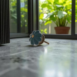 May include: A teal flower-shaped cabinet knob with a gold center. The knob is made of ceramic and has a wooden stem. The knob is resting on a marble surface, with a window and greenery in the background. Decorative hardware.