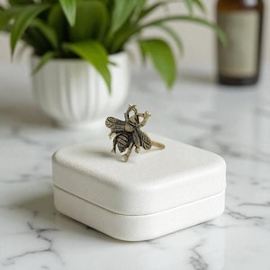 May include: A gold-colored bee ring with intricate detailing, resting on a white square box. The ring's design features a detailed bee with outstretched wings. The background includes a potted plant and a blurred bottle.